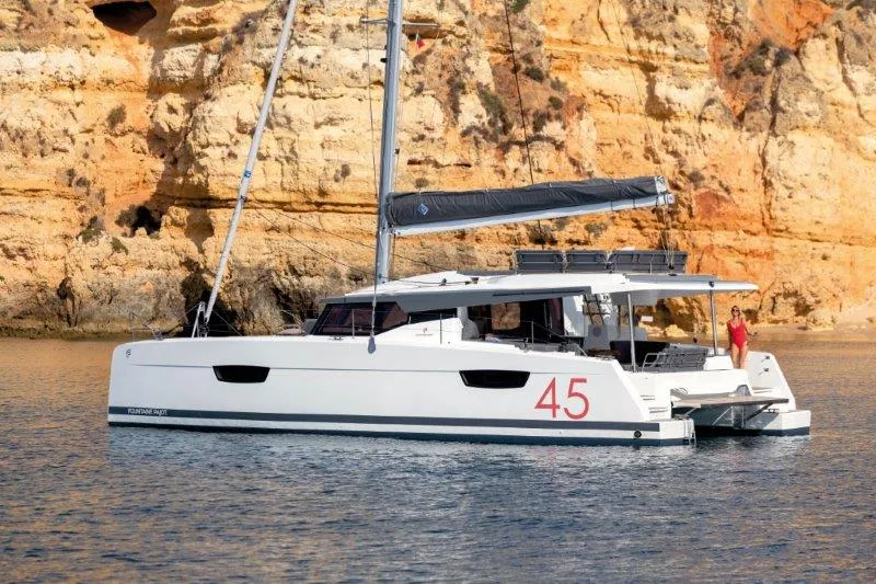 Ein weißer Katamaran Fountaine Pajot Elba 45 schwimmt im Wasser in der Nähe von Klippen.